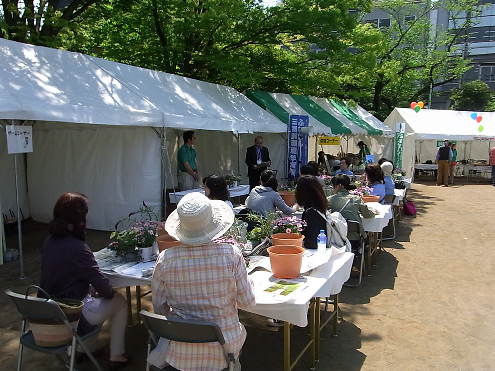 青空の下で約１５人ぐらいの受講生が寄せ植えの仕方を聞いている画像